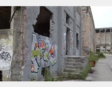 Verlassene industrielle Gebäude aus Beton und Ziegeln mit abbröckelnden Wänden, bemalt mit bunten Graffiti. Vor einem Gebäude führen beschädigte Betonstufen auf einen überwucherten Gehweg.