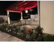 Beleuchtete Terrasse bei Nacht mit einem roten Holzdach und weißen Stützpfeilern, darunter ein Tisch mit weißen Stühlen. Umgeben von einem Blumenbeet mit blühenden Rosen und einer gepflegten Rasenfläche im Hintergrund.