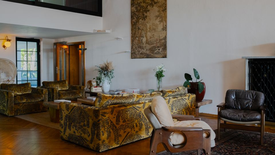 Gemütliches Wohnzimmer mit Sitzmöbeln im Retro-Stil, darunter gelbe Sofas mit schwarzen Mustern und Ledersessel. Der Raum ist mit Holzboden, einem großen Wandteppich, dekorativen Pflanzen und einem Kamin mit Metallverkleidung ausgestattet.