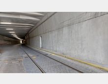Ein Tunnel mit grauen Betonwänden und einer Decke mit sichtbaren Querträgern. Auf dem Boden verlaufen eine Straßenbahnspur sowie ein gepflasterter Fahrweg, beleuchtet durch mehrere Deckenlampen.