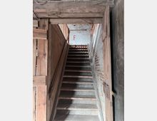 Holztreppe in einem rustikalen Gebäude mit sichtbaren Holzbalken und Backsteinwänden; Tageslicht fällt von oben ein, und an der Wand ist ein rotes "F" sichtbar.