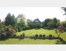 Großer gepflegter Garten mit einer weitläufigen Rasenfläche, umgeben von Bäumen und Sträuchern. Im Hintergrund ist ein großes Haus mit steilem Dach und mehreren Fenstern sichtbar, leicht erhöht hinter einer kleinen Natursteinmauer mit Treppe.