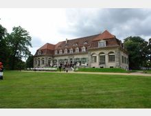 Historisches Herrenhaus mit rotem Mansarddach und großen Sprossenfenstern, umgeben von einer Rasenfläche und Bäumen; im Vordergrund Stühle und Menschen, die vor dem Gebäude stehen.