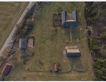 Luftaufnahme eines ländlichen Hofgeländes mit mehreren Gebäuden, darunter Wohn- und Nebengebäude mit schrägen Dächern, umgeben von Wiesen, Feldern und Bäumen. Eine schmale Straße verläuft am linken Bildrand entlang.