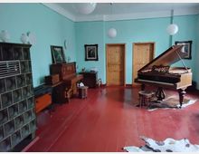 Wohnzimmer mit blauen Wänden und rotem Holzboden, ausgestattet mit einem Flügel, einem Klavier, Möbelstücken im Antik-Stil und einer grünen Kachelheizung. Im Hintergrund befinden sich zwei Holztüren, und die Beleuchtung erfolgt durch hängende Kugelleuchten.