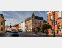 Straßenansicht einer kleinen Altstadt mit historischen Gebäuden, darunter ein großes rotes Backsteinhaus und ein dekoratives Eckgebäude mit spitzem Turm. Kopfsteinpflaster, Laternen und parkende Autos prägen das Straßenbild.