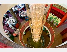 Blick von oben in einen mehrstöckigen, asiatisch gestalteten Innenraum mit roter Dekoration und Verzierungen, einem großen Kronleuchter in der Mitte sowie Dinergästen an runden Tischen auf einem roten Teppichboden. Im unteren Bereich befinden sich große Vasen und dekorative Faltwände mit floralen Mustern.