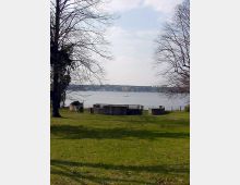 Grüne Parklandschaft mit einem offenen Rasenbereich, großen kahlen Bäumen und einem steinernen Denkmal oder Brunnen in der Mitte. Im Hintergrund ist ein See mit ruhiger Wasserfläche zu sehen, umgeben von einer bewaldeten Uferlinie und vereinzelten Gebäuden.