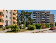 Mehrfamilienhäuser in einer Wohnsiedlung mit vorgelagerten Balkonen in gelben und orangefarbenen Farbtönen, umgeben von Grünflächen und Bäumen. Im Vordergrund befinden sich ein Fußweg, ein Verkehrszeichen, ein Hundekotbeutelspender und ein geparktes Auto.