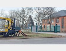 Industriegelände mit gelbem Gleiskran im Vordergrund, einem geschlossenen grünen Tor mit der Beschriftung „Tor 9“ und einem dahinterliegenden Backsteingebäude rechts. Im Hintergrund steht eine kegelförmige, historische Industrieanlage, umgeben von kahlen Bäumen.