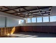 Ein leerer Raum mit Holztäfelung an den Wänden, einer Decke mit Holzbalken sowie eingebauten Deckenleuchten. Große Fenster an der oberen Wand lassen viel Tageslicht herein, der Boden ist mit dunklen Fliesen ausgelegt.