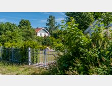 Ein helles Wohnhaus mit rotem Dach liegt hinter dichter Vegetation, umgeben von Bäumen und Sträuchern in einer Gartenlandschaft. Im Vordergrund ist ein Metallzaun mit einem geöffneten Tor sichtbar.