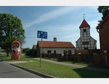 Ländliche Straßenszene mit Fachwerkkirche im Hintergrund, einem Wohnhaus mit rotem Ziegeldach und einem Wegweiser für einen verkehrsberuhigten Bereich; vorne steht eine dekorative Strohpuppe auf einer Wiese.