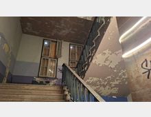 Abgenutztes Treppenhaus in einem älteren Gebäude mit Holzstufen, Metallgeländer und stark abblätternder Farbe an den Wänden. Zwei hohe Fenster mit teils geöffneten Flügeln und abgedeckten Scheiben lassen Licht herein, an den Wänden sind Graffiti zu sehen.