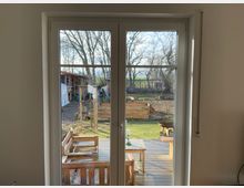 Blick durch eine Glastür auf eine Holzterrasse mit Gartenmöbeln. Dahinter befindet sich ein Garten mit Hochbeeten, Bäumen und einer kleinen Holzhütte.