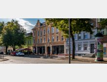 Straßenszene mit historischen Gebäuden, darunter ein orangefarbenes Haus mit der Aufschrift „Flohmarktladen - Möbel aus 2. Hand“, flankiert von weiteren bunt gestrichenen Häusern. Im Vordergrund stehen Autos, ein Baum spendet Schatten, und ein Reklamepfeiler ist sichtbar.
