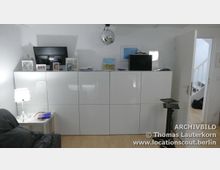 Wohnzimmer mit weißer Hochglanz-Kommode, auf der diverse Dekorationsgegenstände wie ein Monitor, Bilderrahmen und eine Lampe stehen. Rechts führt eine offene Tür zu einer Treppe, links steht ein kleiner Beistelltisch mit einer Lampe und darauf liegenden Büchern neben einem Sofa.