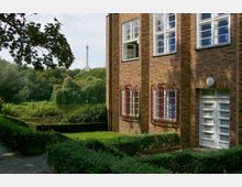 Seitenansicht eines Backsteingebäudes mit weißen Fenstern und roten Fenstergittern, umgeben von gepflegten Hecken und einem Garten; im Hintergrund ein Teich mit Weidenbäumen und der Berliner Funkturm im Dunst sichtbar.