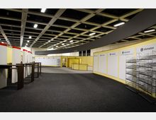 Ein leerer Messe- oder Konferenzbereich mit gelben Wänden und Informationstafeln, leeren Regalen und Schreibtischen aus Holz. Die Decke besteht aus offenen Gitterstrukturen mit eingebauten Leuchtstofflampen.