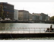 Blick auf eine Uferpromenade mit mehrstöckigen Gebäuden im Hintergrund, darunter Baustellen und ältere Fassaden. Im Vordergrund ein Wasserbereich mit einer abgesperrten, niedrigen Plattform, auf der Sonne reflektiert wird.