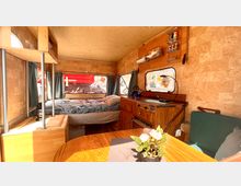 Innenansicht eines Wohnwagens mit Holzausbau: Im Vordergrund ein Esstisch mit einer Topfpflanze, rechts eine kleine Sitzbank, links ein Bett mit blauer Bettwäsche. Im Hintergrund eine Küchenzeile mit Gasherd, Hängeregalen und einem Fenster mit Vorhängen.