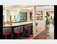 Eine moderne Wohnküche mit einer langen Kücheninsel aus hellem Stein, umgeben von gepolsterten Barhockern mit gemusterten Bezügen und roten Sitzen. Im Hintergrund sind grüne Schränke, offene Regale und Pflanzen auf Holz- und Fliesenböden zu sehen.