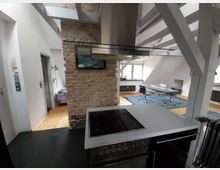 Wohnbereich mit offener Küche in einem Dachgeschossraum. Sichtbare Holzbalken, eine freistehende Ziegelwand, ein moderner Herd mit Dunstabzugshaube, ein angrenzender Essbereich mit Glas-Esstisch und Stühlen sowie ein Holzboden und Dachschrägen prägen den Raum.