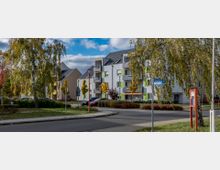 Wohnsiedlung mit mehrstöckigen, modernen Gebäuden in hellen Farben und grünen Akzenten, umgeben von herbstlichen Bäumen und Sträuchern. Im Vordergrund befindet sich eine Straße mit einem Wegweiser „Karlplatz“ und einem roten Informationsschild.