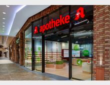 Moderne Apotheke in einer Passage mit Backsteinfassade; durch die Glasfront sind Regale mit Produkten sowie runde Präsentationstische sichtbar, beleuchtet von Deckenleuchten. Ein rotes Apotheken-Logo und die Beschriftung „apotheke“ befinden sich über dem Eingang.