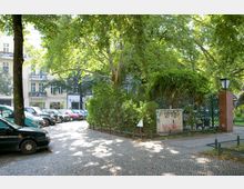 Kleiner städtischer Platz mit Kopfsteinpflaster, parkenden Autos und großen schattenspendenden Bäumen. Im Hintergrund ein mit Efeu bewachsener Zaun, eine rote Backsteinsäule mit einer Laterne und ein Stromkasten mit Graffiti.