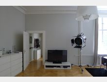 Wohnzimmer in einer Altbauwohnung mit hellgrauen Wänden und Stuckdecke. Zu sehen sind ein Fernseher auf einem weißen TV-Möbel, ein großes Filmlicht auf einem Stativ, eine helle Tür, die in einen Flur führt, sowie ein großes Fenster mit beigefarbenen Vorhängen. Der Boden besteht aus hellem Parkett.