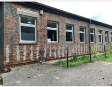 Backsteinfassade eines einstöckigen Gebäudes mit weißen Fenstern in regelmäßiger Anordnung. Der Bereich vor dem Gebäude zeigt einen gepflasterten Weg, eine grüne Rasenfläche und ein Zaunelement.