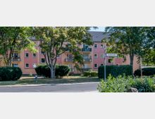 Mehrstöckiges Wohnhaus mit rosa Fassade, rechteckigen Fenstern und Balkonen, umgeben von Bäumen und Sträuchern. Im Vordergrund ist eine Straßenkreuzung mit einem Straßenschild sichtbar.