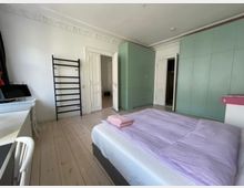 Ein helles Schlafzimmer mit einem Doppelbett, das mit lila Bettwäsche bezogen ist. An der linken Wand befinden sich ein weißer Schreibtisch, ein schwarzes Wandregal und eine geöffnete Tür, während die rechte Wand von türkisfarbenen Einbauschränken dominiert wird. Der Boden ist aus hellem Holz, und die Decke ist mit Stuckornamenten verziert.