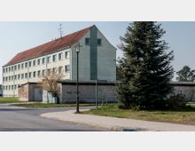 Ein mehrstöckiges Wohngebäude mit blassgrüner Fassade und rotem Satteldach steht neben einer flachen Garagenstruktur. Im Vordergrund ist ein Gehweg mit einer Straßenlaterne, einer Tanne und einer Straßennamensschild mit der Aufschrift "Kameruner Weg" zu sehen.