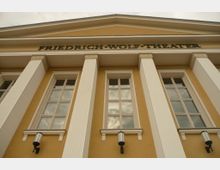 Außenansicht des Friedrich-Wolf-Theaters, gekennzeichnet durch gelbe Fassade, weiße Säulen mit klassischen Verzierungen und rechteckige Fenster sowie Wandlaternen. Über dem Eingangsbereich ist der Name des Theaters in großen Buchstaben angebracht.