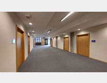 Ein breiter, heller Korridor mit grauem Teppichboden, weißen Wänden und mehreren Holztüren auf beiden Seiten. Am Ende des Flurs befindet sich eine Glastür mit einem großen Fenster daneben, das Tageslicht hereinlässt; dahinter ist eine Backsteinwand sichtbar.