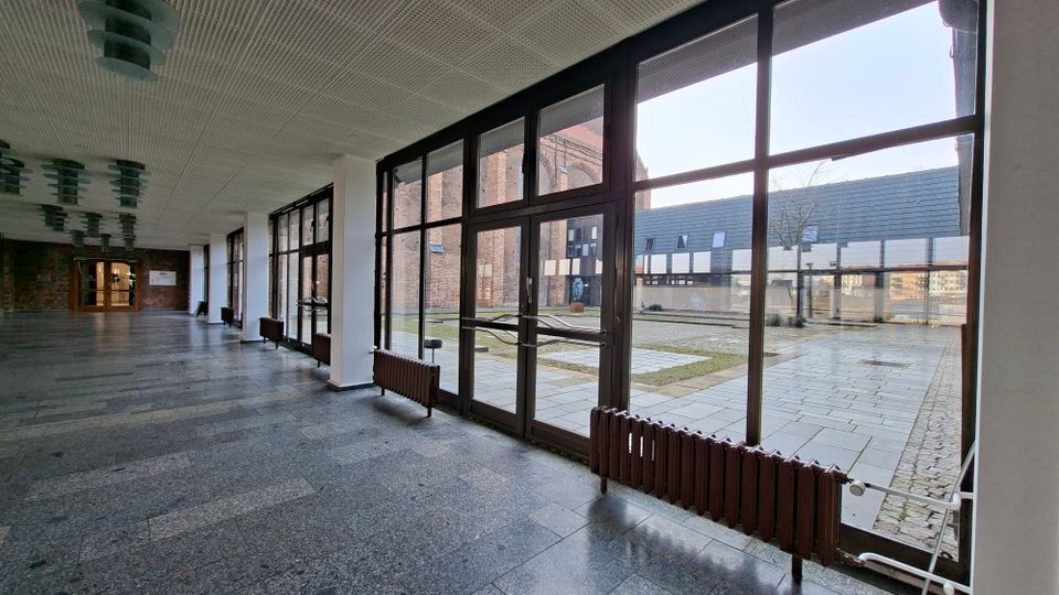 Langer, überdachter Gebäudeflur mit dunklem Steinboden und einer durchgehenden Reihe großer, schwarz gerahmter Glasfenster und -türen. Durch die Fenster ist ein gepflasterter Innenhof mit niedrigen Gebäuden und einer Backsteinfassade zu sehen.