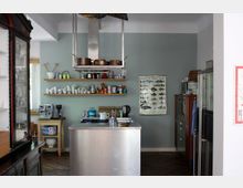 Moderne Küche mit zentraler Kücheninsel aus Edelstahl, einer Dunstabzugshaube und Holzregalen an der Wand, die Geschirr, Tassen und Vorratsbehälter präsentieren. Im Hintergrund ist eine Wand mit Fischposter und eine Kaffeemaschine auf einem kleinen Holzschrank sichtbar.