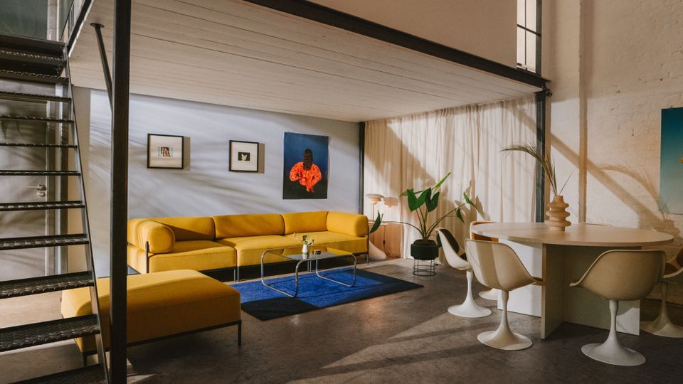 Modernes Loft-Wohnzimmer mit Zwischenebene und schwarzer Metalltreppe, Betonboden und hoher Fensterfront mit Vorhängen. Gelbes Ecksofa mit Glastisch auf blauem Teppich neben einem runden Esstisch mit weißen Tulpenstühlen, dazu Wandkunst und Zimmerpflanzen.