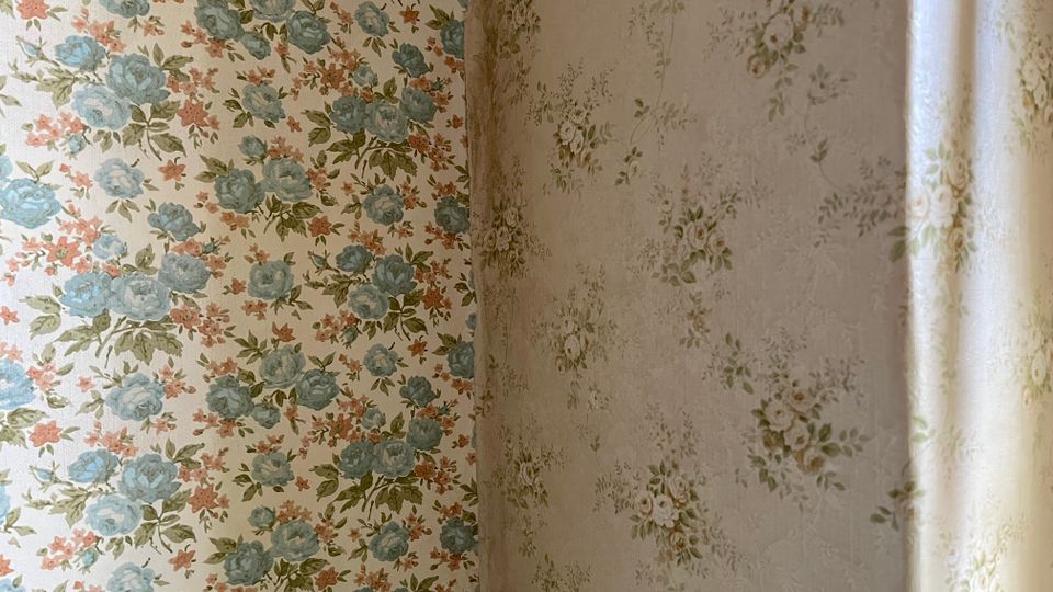 Nahaufnahme einer Raumecke mit zwei unterschiedlich gemusterten Blumentapeten, links mit blau-rosa Blumen und rechts mit dezenteren, kleinen Blumenmotiven. Rechts fällt weiches Tageslicht entlang der Wand in den Raum.