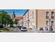 Straßenszene in einer Kleinstadt mit Pflastersteinstraße, geparkten Autos und beigefarbenen Wohngebäuden im Vordergrund. Im Hintergrund ist ein Kirchturm mit Ziffernblatt und spitzer Turmspitze sowie rote Ziegeldächer zu sehen.