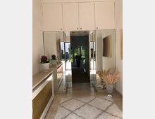 Langer Flur mit beigefarbenem Fliesenboden, einer flauschigen Teppichläufer in der Mitte und großen, wandhohen Spiegeln an den Wänden. Rechts und links stehen Dekorationselemente wie Pflanzen und Trockenblumen, im Hintergrund ist durch eine Glastür ein Garten sichtbar.