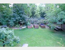 Grüne Gartenanlage mit Rasenfläche, mehreren Hochbeeten aus Holz, einem kleinen Tisch mit Stühlen sowie einer Sitzbank und einem Picknicktisch im Hintergrund. Die Umgebung ist von Bäumen und dichter Vegetation umgeben, mit einem Holzzaun am Rand.