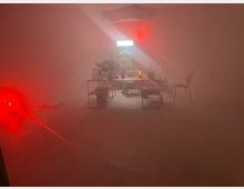 Ein industriell gestalteter Raum mit minimaler Ausstattung, schwacher Beleuchtung und rotem Nebeleffekt. In der Mitte stehen durchsichtige Tische mit Gegenständen darauf, daneben befinden sich vereinzelt Stühle; im Hintergrund ist ein Regal mit Flaschen unter kaltem Licht sichtbar.