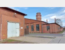 Backstein-Industriegebäude mit einem hohen, runden Turm im Hintergrund. Die Fassade ist geprägt von großen Fenstern mit Sprossen, einer großen Metalltür und einem schlichten Schild mit der Aufschrift „Gebäude 14“.