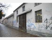 Graffiti-bemalte Hausfassade entlang einer gepflasterten Gasse, mit vergitterten Fenstern und einem großen Holztor; im Hintergrund kahle Äste und weitere Gebäude.