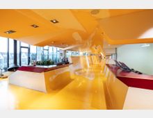 Modernes, farbenfrohes Innenraumdesign mit einer geschwungenen, gelb-weißen Decken- und Bodenstruktur, die sich durch den Raum zieht. Große Fenster lassen Tageslicht herein, und ein dunkelroter Empfangstisch mit Monitoren dominiert den Vordergrund.