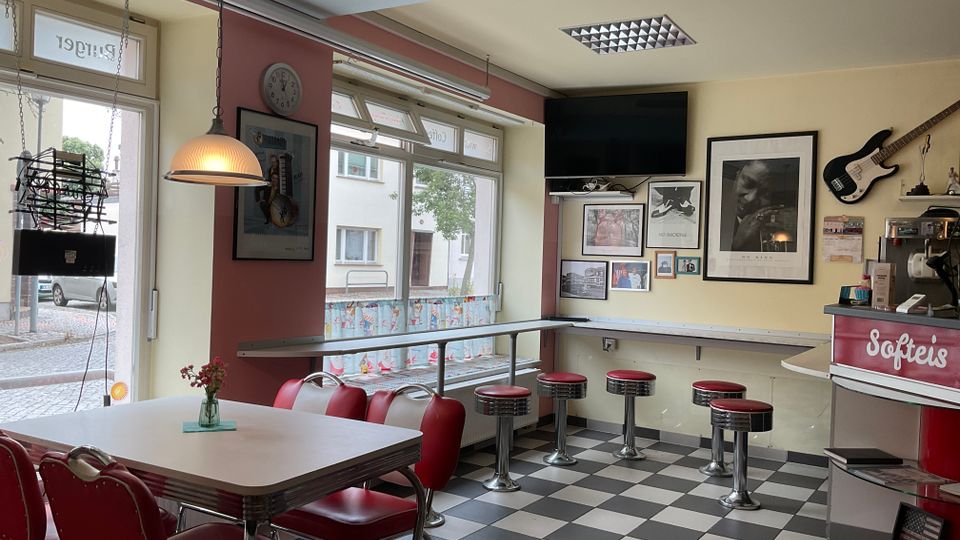 Innenansicht eines kleinen Diners mit schwarz-weiß kariertem Boden, roten Sitzbänken und Chrom-Barhockern an einer Fensterbar. An der Wand hängen gerahmte Bilder und eine E-Gitarre, daneben steht eine rote Theke mit der Aufschrift „Softeis“ und großen Fenstern zur Straße.