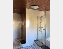 Badezimmer mit einer Dusche mit Glasabtrennung, weißen Fliesenwänden und einem Holzpaneeldeckenelement, daneben ein Bereich mit Sauna und einer kleinen gefliesten Sitzfläche. Der Boden ist gefliest im schwarz-weißen Mosaikmuster.